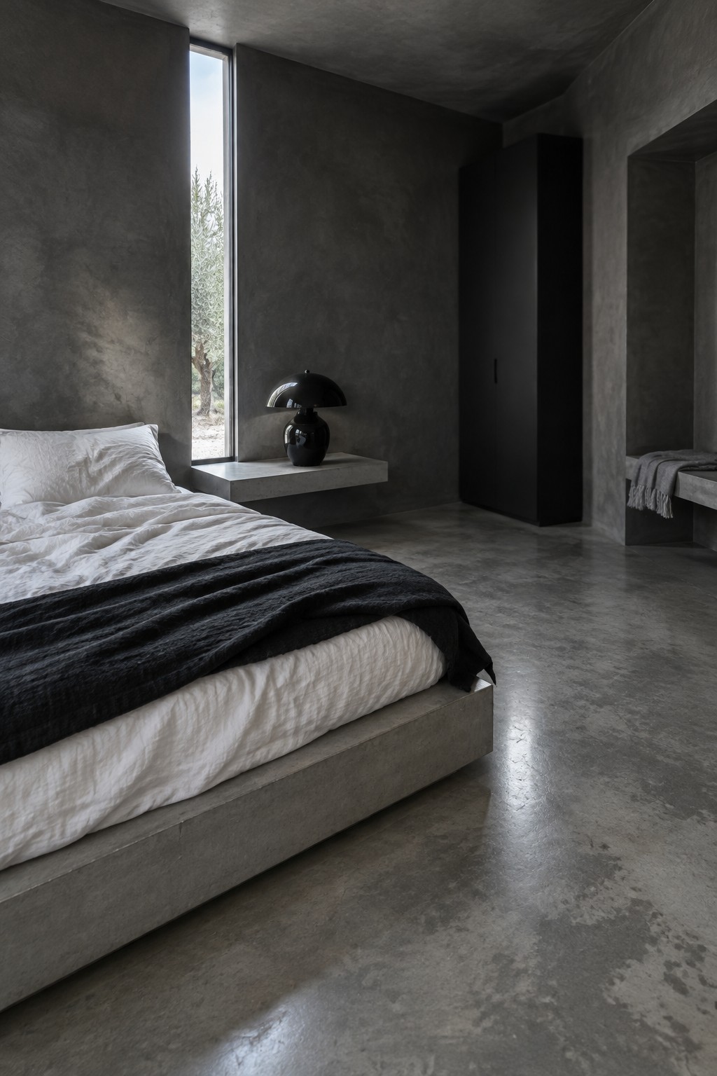 A bedroom with charcoal concrete walls, a low concrete bed, white bedding, and a narrow vertical window.